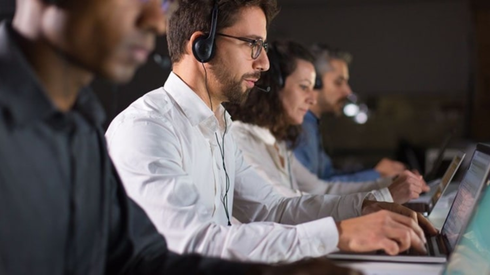 Side view of confident call center operator talking with client. Caucasian young man in eyeglasses typing on laptop while serving client. Call center concept